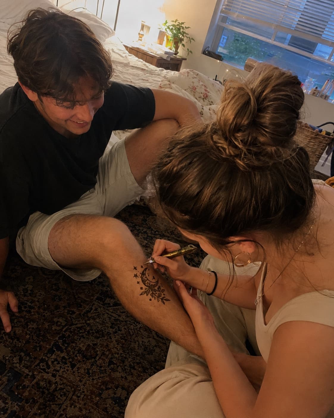 Carefully inking a henna moon design on a partner's knee, sitting on the floor by the bed