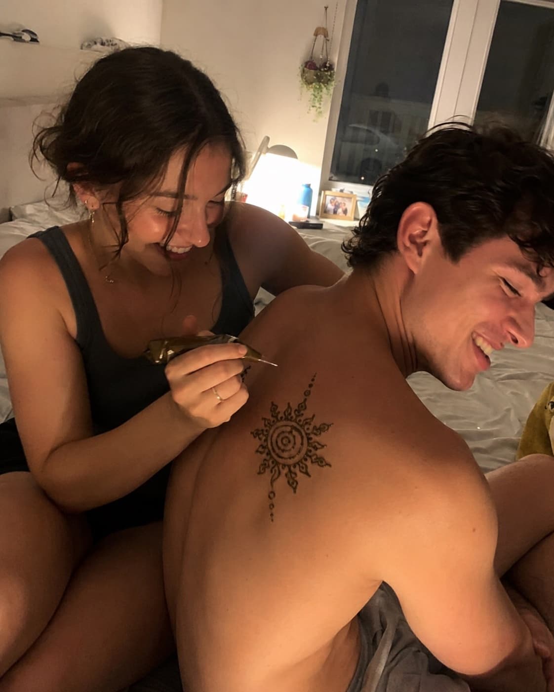 One partner drawing a henna sun on the other's shoulder blade, both laughing on the bed at home